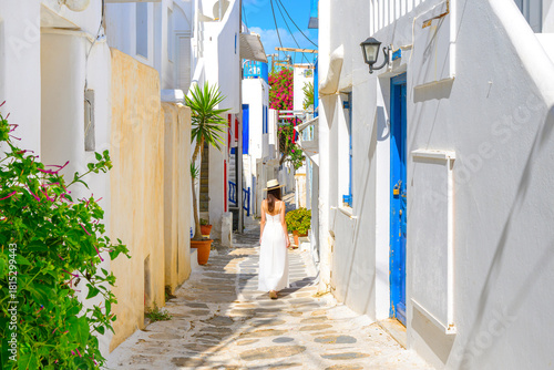 Fototapeta Naklejka Na Ścianę i Meble -  A young woman walks down a narrow whitewashed street with colorful doorways and bougainvillea flowers in the old town center of Mykonos, Greece.