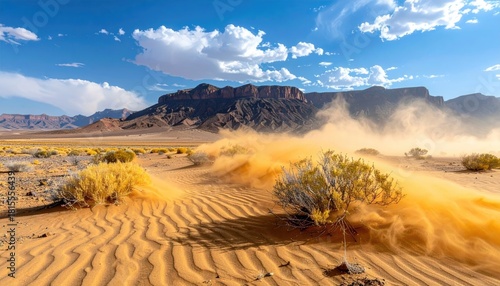 Fototapeta Naklejka Na Ścianę i Meble -  Desert landscape with wind blowing sand across textured dunes under a bright blue sky with scattered clouds and arid vegetation in the distance.