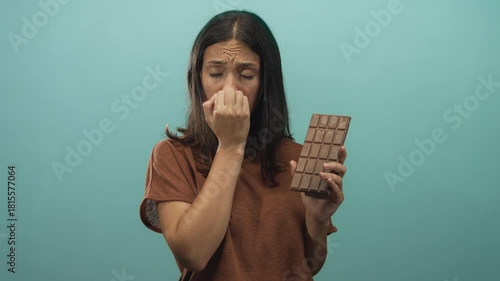 Young hispanic woman biting nails and holding a large chocolate bar in studio with teal backdrop; anxiety craving temptation.