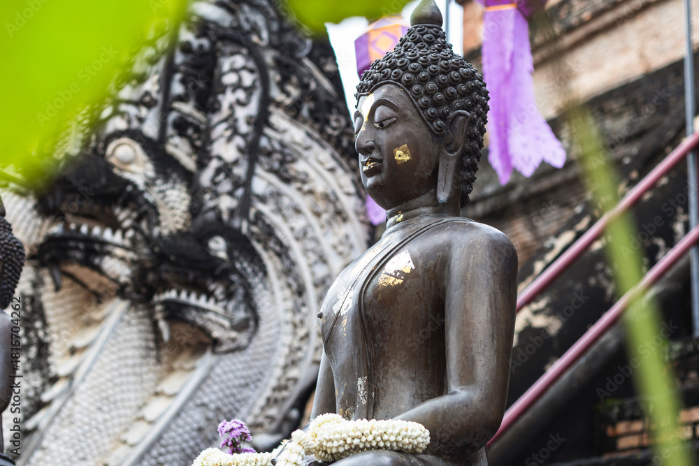 Obraz premium Serene bronze Buddha with gold leaf, jasmine garlands sits peacefully at a Chiang Mai temple. Blurred Naga sculptures, purple lanterns in background.