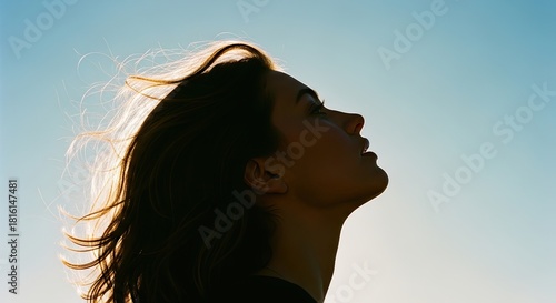 A serene woman's face in profile looking up towards the light with a hopeful expression, her hair blowing in the wind and backlit by the beautiful morning sun