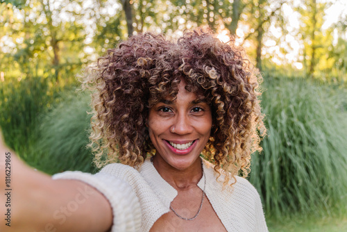 Foto Smiling young woman with curly highlighted hair taking a selfie in a green park