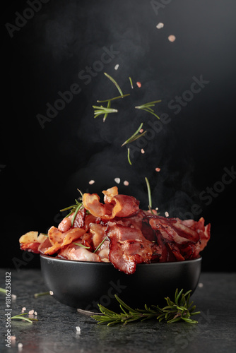 Fried bacon in a black dish, sprinkled with rosemary and pink salt.