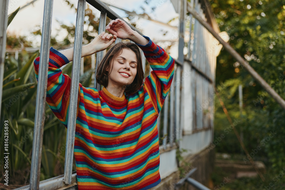 Obraz premium Woman in colorful sweater with smile, relaxed in greenhouse, portrait with closed eyes enjoying plants and natural light. Leisure, cozy knit, autumn mood and serene expression.