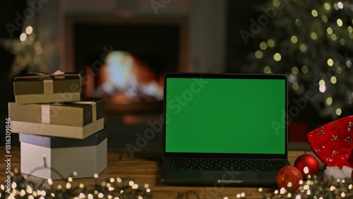Laptop computer, presents, gift boxes at Christmas. Decorated room with Christmas tree and lights. Cosy winter home with fireplace. Blank green screen for web shop offer and holiday advertisement.
