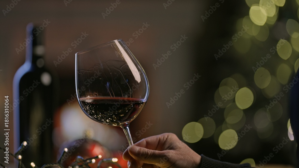 Fototapeta premium Glass and bottle of red wine on table in Christmas decorated home. Man swirling tasting buying wine. Photo for web shop ads and vinery holiday offer.