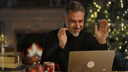 Virtual Christmas online - Happy smiling middle aged man hat at home video chatting with family on laptop computer, talking, waving hand. Cosy room with fireplace in winter.