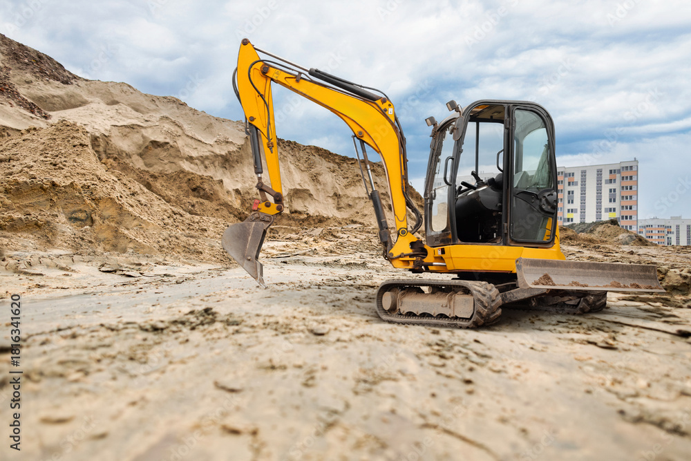 Obraz premium An excavator works to clear sand at a construction site surrounded by tall buildings under a cloudy sky, showcasing urban development activities