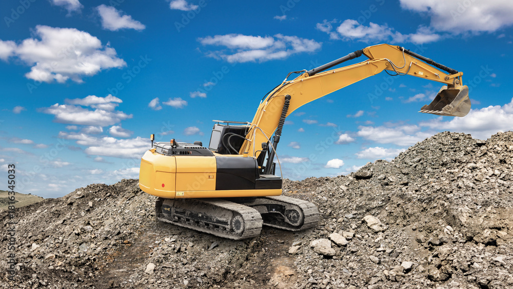 Obraz premium An excavator is moving dirt at a construction site with a clear blue sky and scattered clouds overhead. Piles of gravel and rocks are visible around the machine