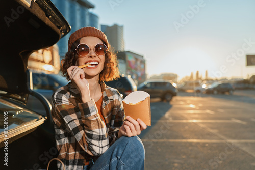 Photography woman eats fries by car trunk in parking lot, smile and sunglasses catching sunl
