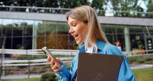Modern female professional in casual clothes holding laptop and using phone while walking in business district park