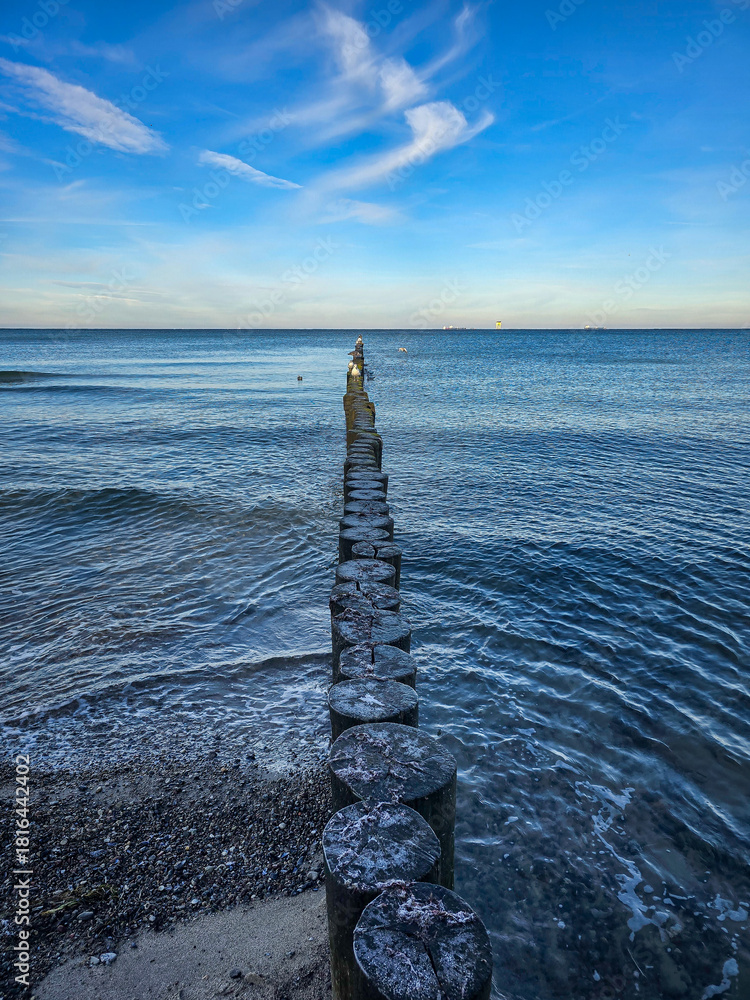 Fototapeta premium Weathered wooden groynes stretch into the gently rippling sea beneath a blue sky streaked with clouds, forming a peaceful coastal scene at the edge of a pebbled beach