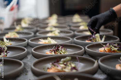Professional kitchen staff meticulously plate dozens of gourmet appetizers, adding delicate microgreens and herbs for an upscale catering event.