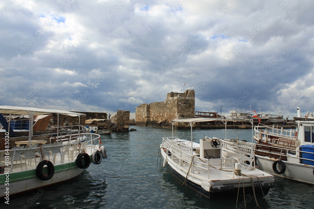 Obraz premium White tour boats docked at historic stone harbor near ancient fortress under dramatic cloudy sky.