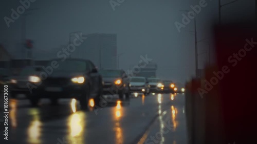 Vehicles drive slowly along a wet highway during a heavy rainstorm in the evening. Reflections of lights can be seen on the road, creating a hazy and moody atmosphere