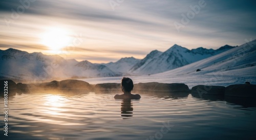 Person relaxing in steaming outdoor mineral bath surrounded by snowy mountain landscape during sunrise or sunset