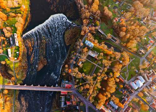 Aerial top down view of small European town with river waterfall, bridge, autumn trees and colorful houses on a sunny evening, scenic urban landscape background