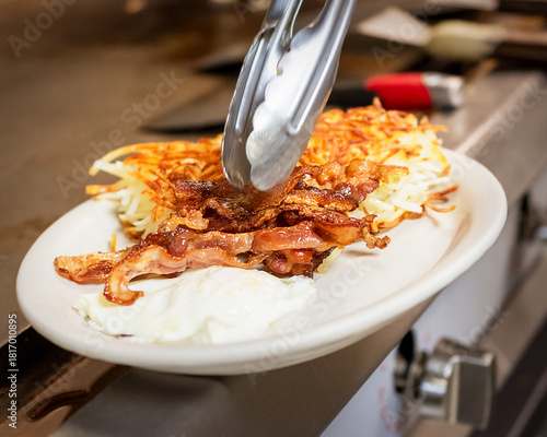 Obraz na plátně A chef uses tongs to place crispy bacon on a plate of fried eggs and golden hash browns, completing a classic cafe breakfast