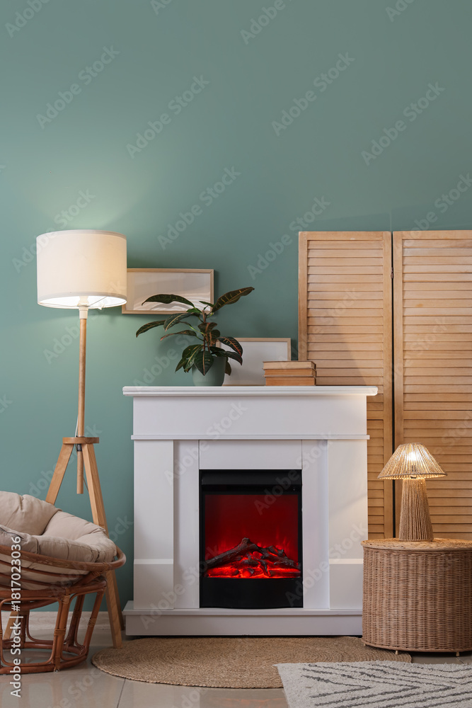 Fototapeta premium Interior of living room with folding screen, glowing lamps, armchair, wicker table and home decor on fireplace near blue wall