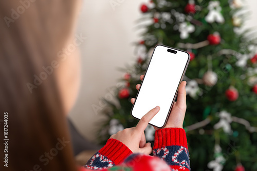 Girl holding smartphone with blank screen near Christmas tree, perfect for holiday app mockups, seasonal advertising, mobile promotions and festive digital designs