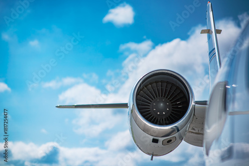 The jet engine and tail fin of a small private jet. The focus is on the engine fan and cowling. A blue sky filled with clouds is in the background.