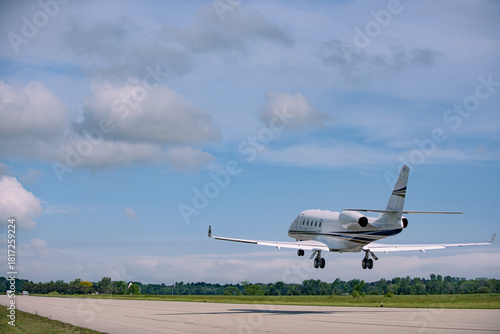 Rear view of a Gulfstream 260 beginning climb-out, wheels off the runway, wide angle view showing early ascent under bright daylight with soft clouds and tree line.