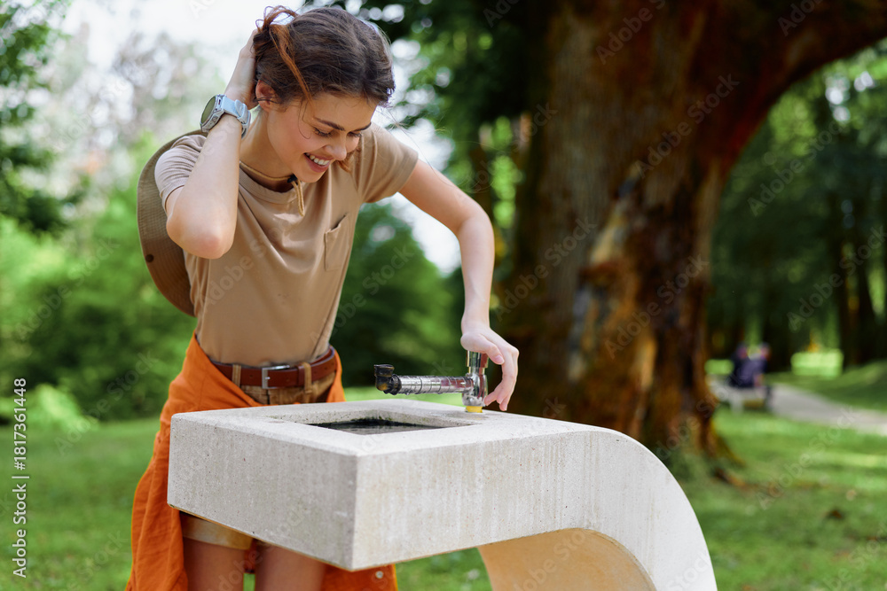 Naklejka premium Woman drinking water from a public fountain in a green park, pouring bottle into basin while smiling, casual summer outfit, outdoor leisure and hydration moment.