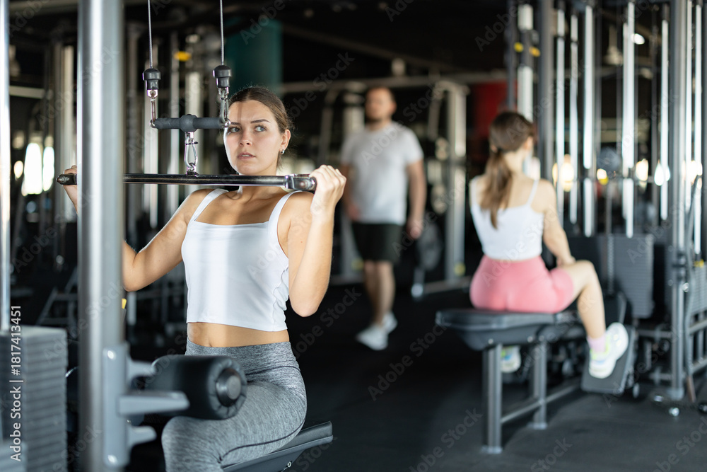 Fototapeta premium Young athletic woman training her arms using machine in gym