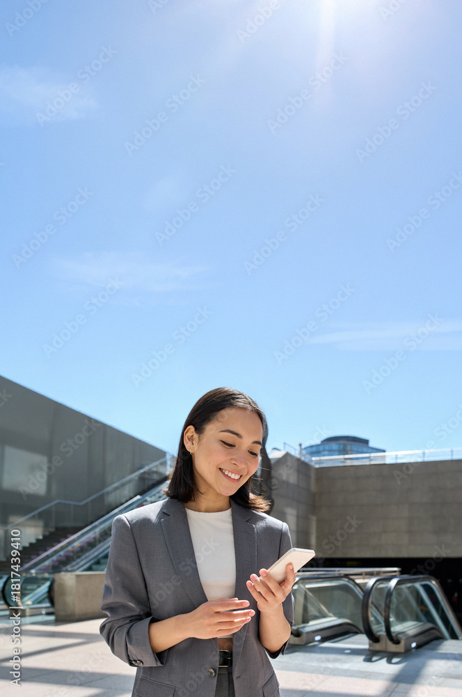 Naklejka premium Young happy Asian business woman wearing suit holding mobile phone standing in city subway using smartphone for texting, checking apps for public transport, metro or travel guide