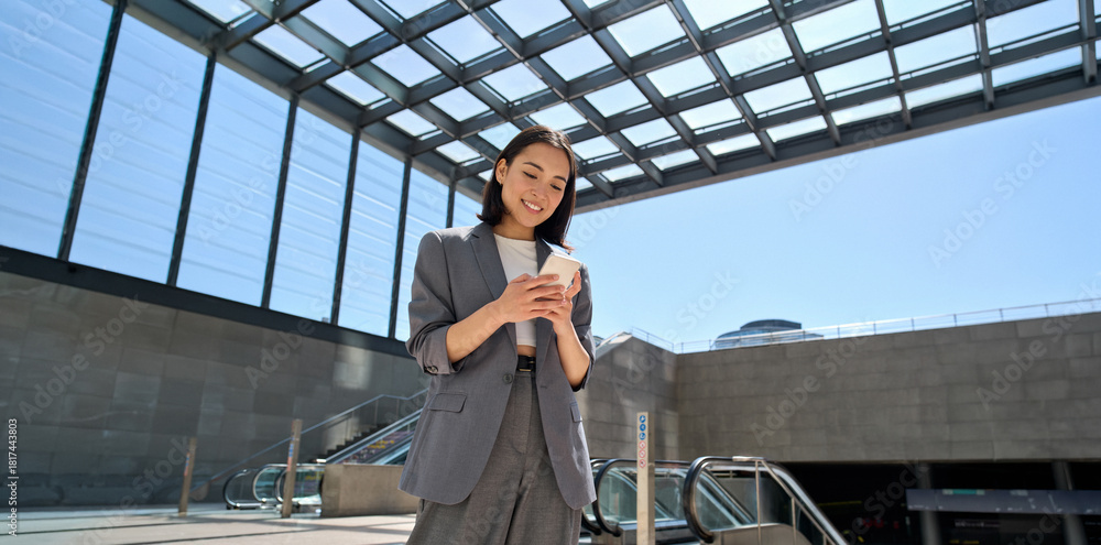 Naklejka premium Young smiling Asian business woman wearing suit holding cell phone standing in city metro using smartphone fast connection, checking apps for public transport or travel navigation maps guide