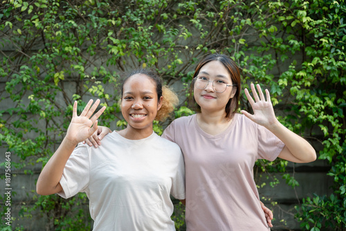 Happy friends waving and smiling outdoors, Two young women posing together and greeting in garden, Cheerful diverse friends standing arm in arm and waving at camera