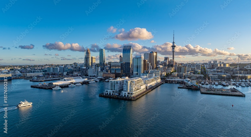 Naklejka premium Auckland, New Zealand cityscape at sunset. A boat cruises on the water in the foreground, with the city skyline and Sky Tower in the background. The photo captures the beauty of the city.