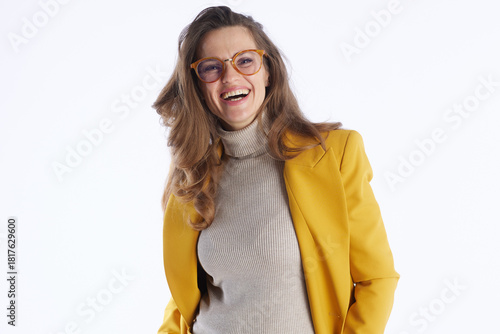 A cheerful professional woman with wavy brown hair and glasses smiles confidently in a bright yellow blazer and beige turtleneck against a clean white background.