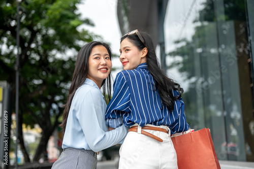 Pretty asian woman friends with shopping bag linking arm turning looking back walking outside a mall