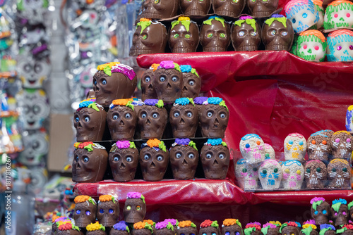 A display of chocolate skulls with flowers on them. The skulls are arranged in a pyramid shape. Mexican Day of the Dead tradition, offerings, food, colors, decorations, and papel picado, La Catrina.