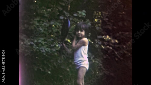 Little girl playing badminton with racket in lush green backyard, embodying innocence and carefree fun. Vintage footage capturing childhood moment. Old film. Archival retro 1960s. Aged archive.