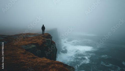 A solitary person standing on the edge of a rugged cliff overlooking a misty ocean with crashing waves under a foggy sky during daytime