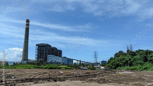 Industrial landscape featuring power plant with towering chimney against bright blue sky generating energy and fuel for cities and homes