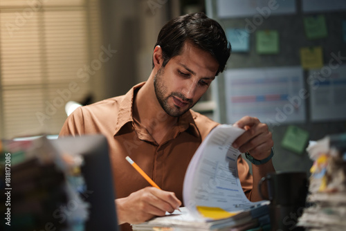 Latin man reviews financial accounting documents and tax paperwork, checking each report and file in an office at night