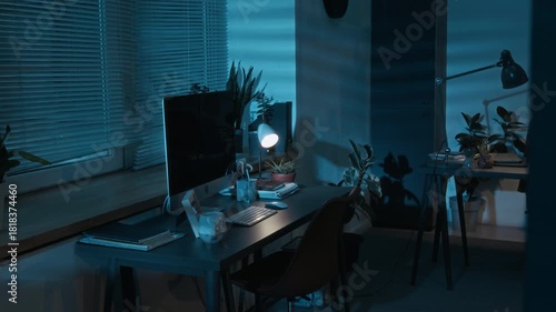 Dark empty office showing window with blinds and grey work table with computer monitor, wireless keyboard, notebooks, decorative objects and lighted lamp