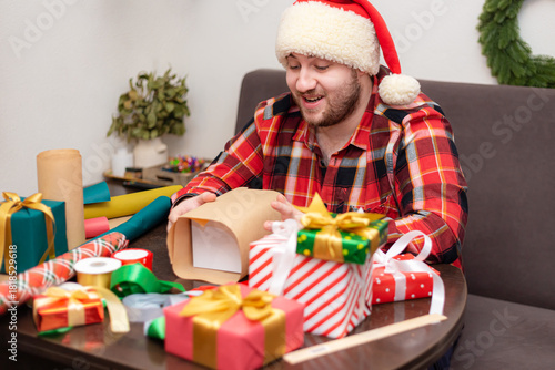 A young, bearded Caucasian man wearing a plaid shirt wraps Christmas presents.