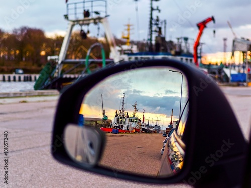Harbor view reflected in a car side mirror at sunset, showing fishing boats and industrial port details.