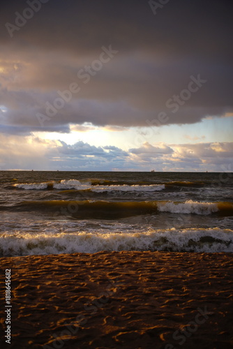 Fototapeta Naklejka Na Ścianę i Meble -  The sunset time golden hour walk to the Baltic sea in Lithuania, Europe, evening view of the yellow and red clouds, ocean waves in storm, girl walking in sport shoes, grey sand grains travel lifestyle