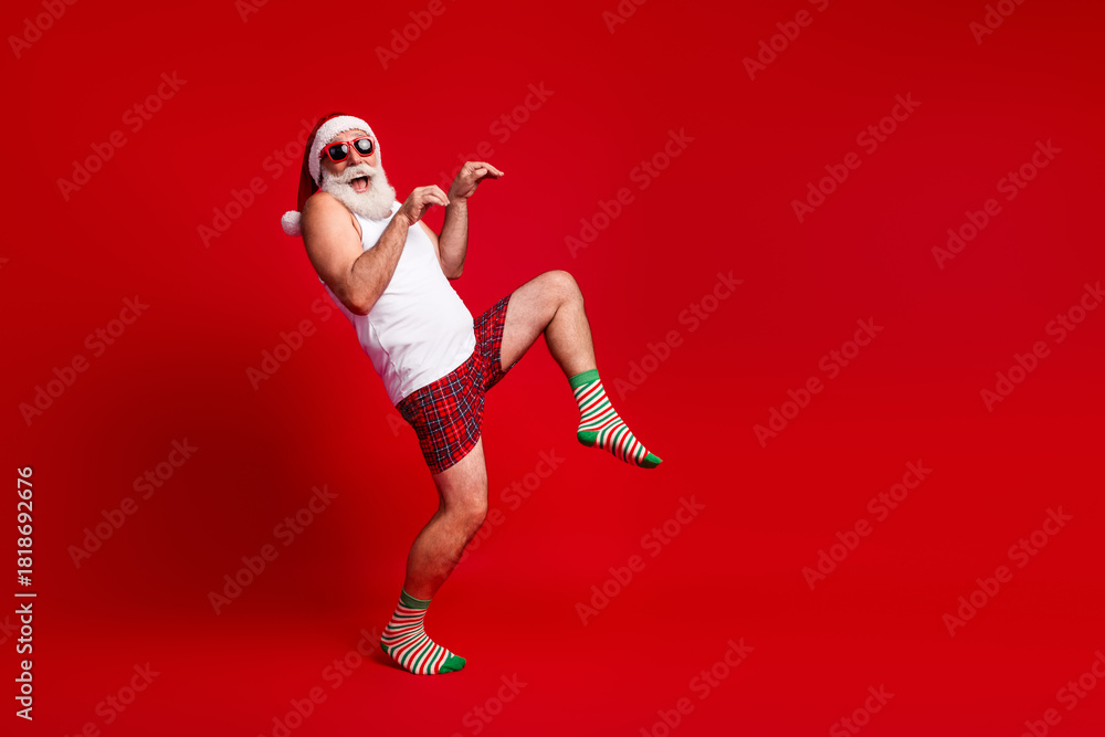 Obraz premium Playful Santa look alike dances in bright red studio wearing white tank top plaid shorts and striped socks for festive christmas vibe