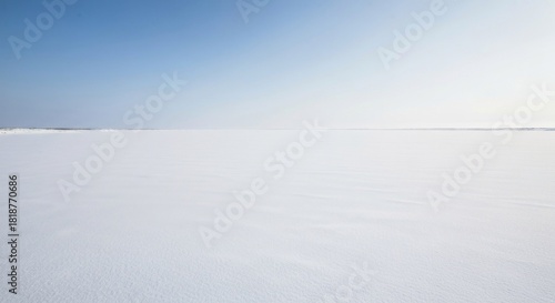Vast, pristine expanse of snow-covered ground under a clear, light-blue sky. Gentle, horizontal lines suggest slight wind patterns across the surface. Horizon is indistinct