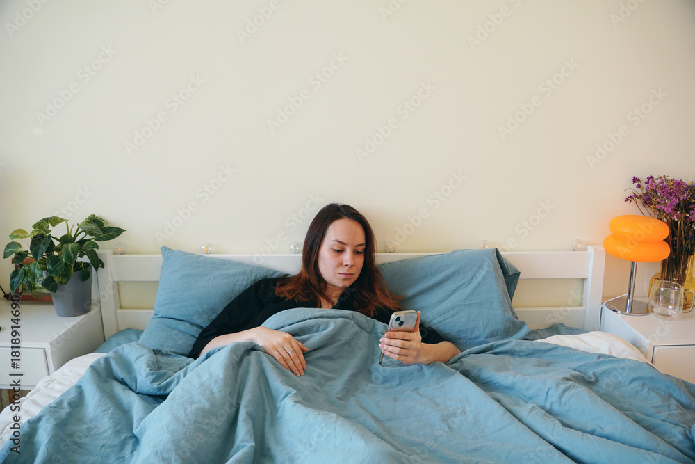 Obraz premium A young woman lies in bed in a cozy bedroom and looks at her phone. The girl checks her email and social media after waking up