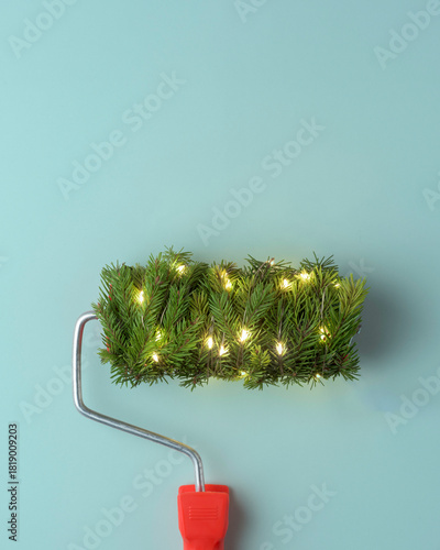A festive paint roller covered in bright evergreen branches and warm lights creates a playful holiday mood on a soft pastel background.