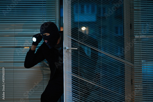 Принти на полотні Man wearing black balaclava entering office at night holding flashlight, crouchi