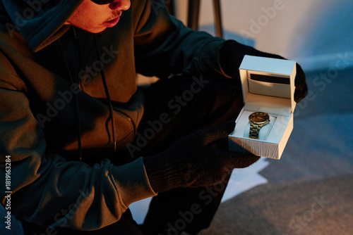 Young adult man wearing gloves and hoodie holding open watch box with gold wristwatch inside, crouching in office environment, engaging in theft activity