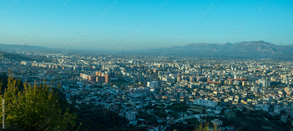 Obraz premium A sweeping panoramic view of the vast Tirana cityscape extending toward the horizon, backed by a series of low mountains under a hazy, bright blue sky.